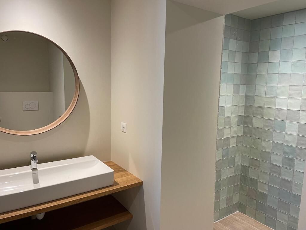 A bathroom with a rectangular sink, a round mirror, and a tiled shower wall.