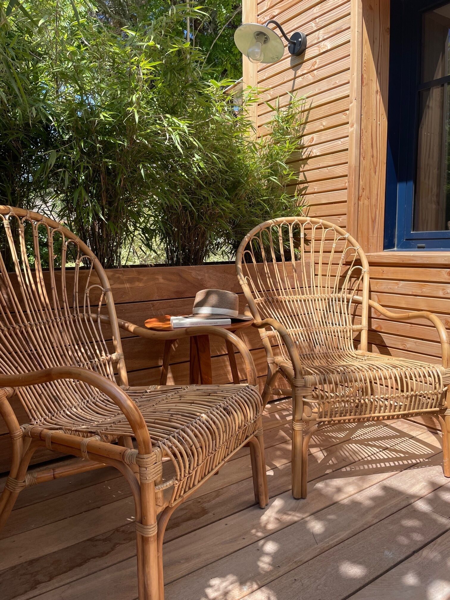 Une terrasse en bois avec deux fauteuils en osier et une table, un luminaire fixé au mur et une fenêtre.