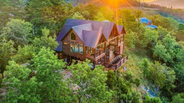A large wooden cabin with multiple levels, surrounded by dense forest, and featuring a prominent deck area. - Breathtaking Views, Hot Tub (Broken Bow)