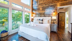 A bedroom with a large bed, wooden walls, and a view of trees through large windows. - Breathtaking Views, Hot Tub (Broken Bow)