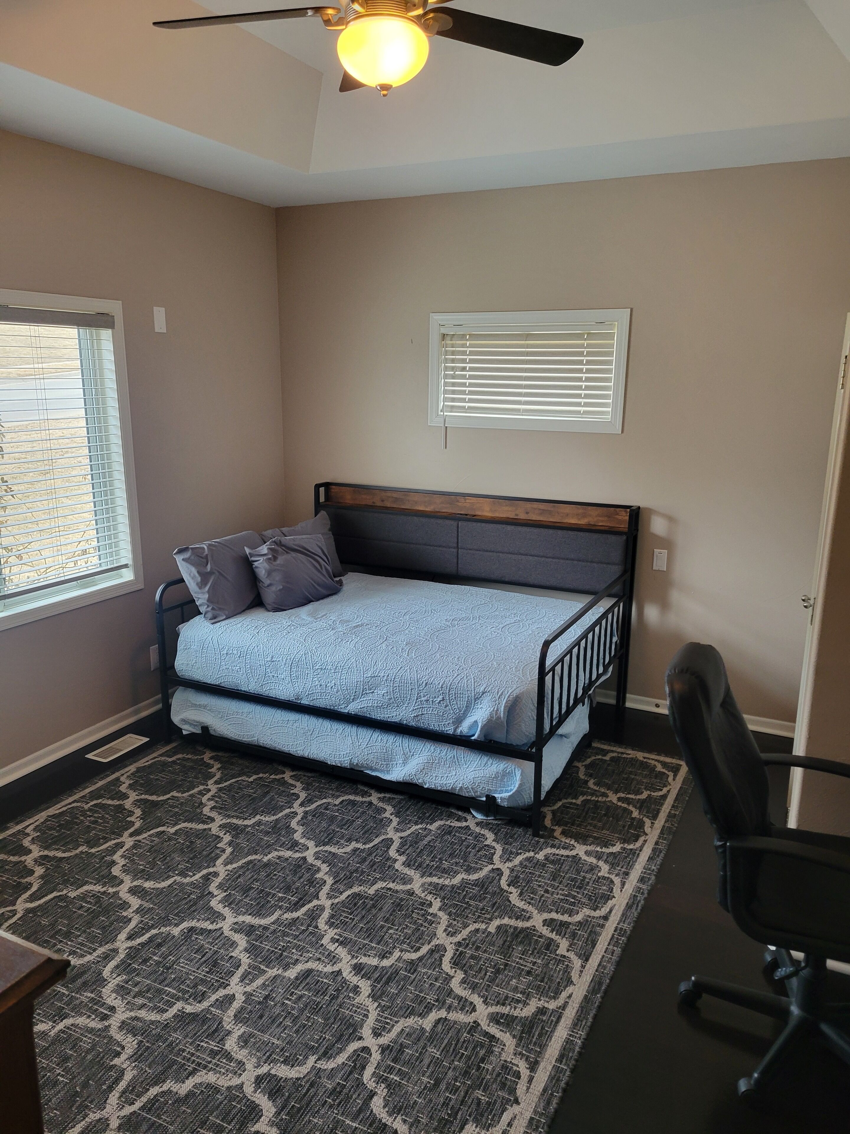 A bedroom with a bed, a ceiling fan, a window with blinds, a chair, and a patterned rug.