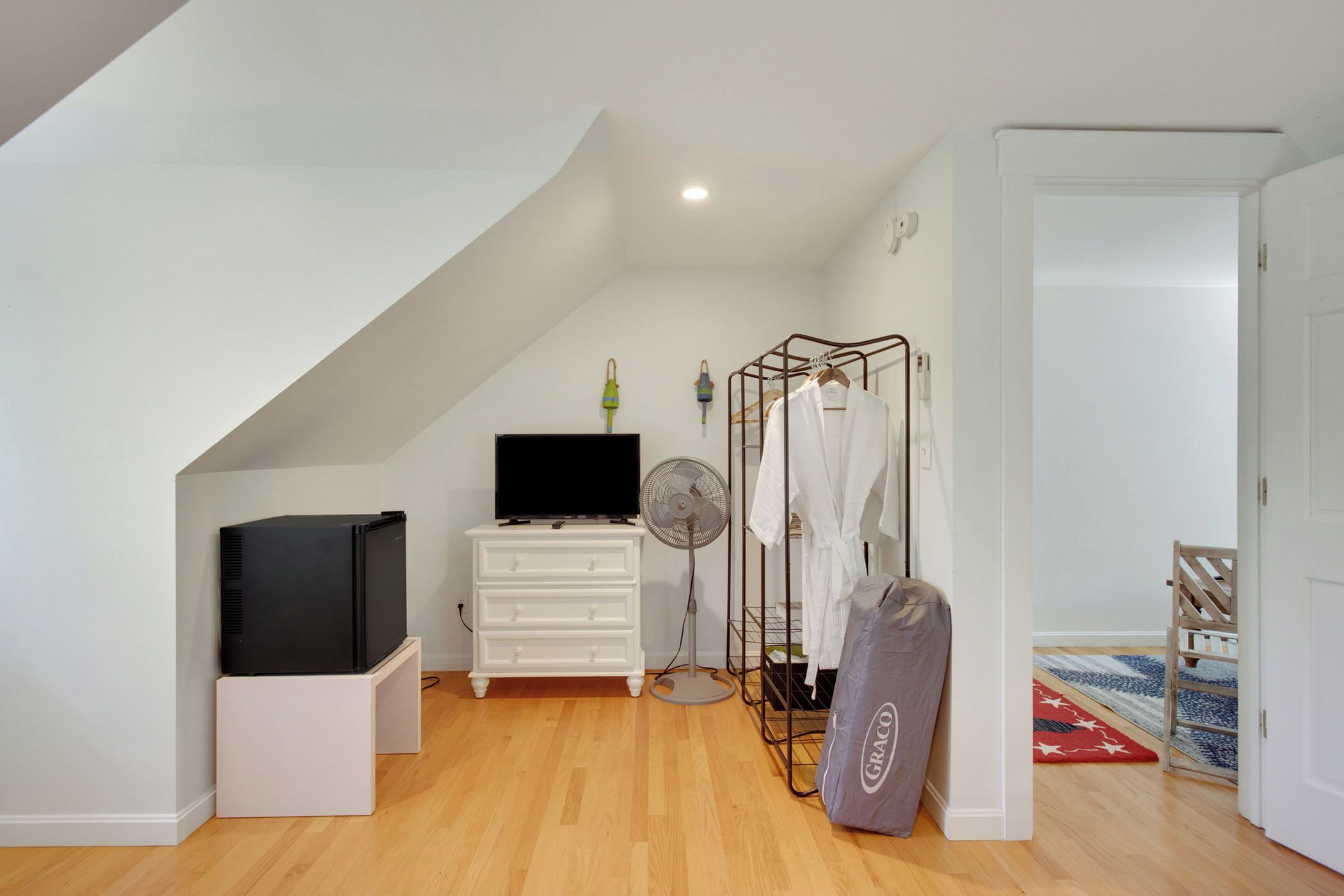 A room with a TV on a stand, a white dresser, a fan, a coat rack with clothes, and a door leading to another room.