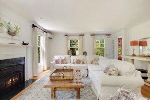Uma sala de estar com lareira, sofás brancos, uma mesa de centro e uma janela grande com cortinas.