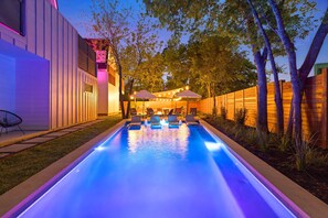 Piscina con sillas de descanso y sombrillas, rodeada por una valla de madera y árboles.