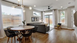 A modern living room with a round wooden dining table, black wireframe chairs, a grey sofa, and a ceiling fan. - InstaWorthy Group Travel Retreat | 3.5 Miles to DT (Austin)