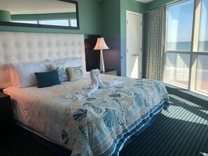 Ein Hotelzimmer mit einem großen Bett, einem Nachttisch, einer Lampe, einem Kleiderschrank und Meerblick durch ein Fenster.