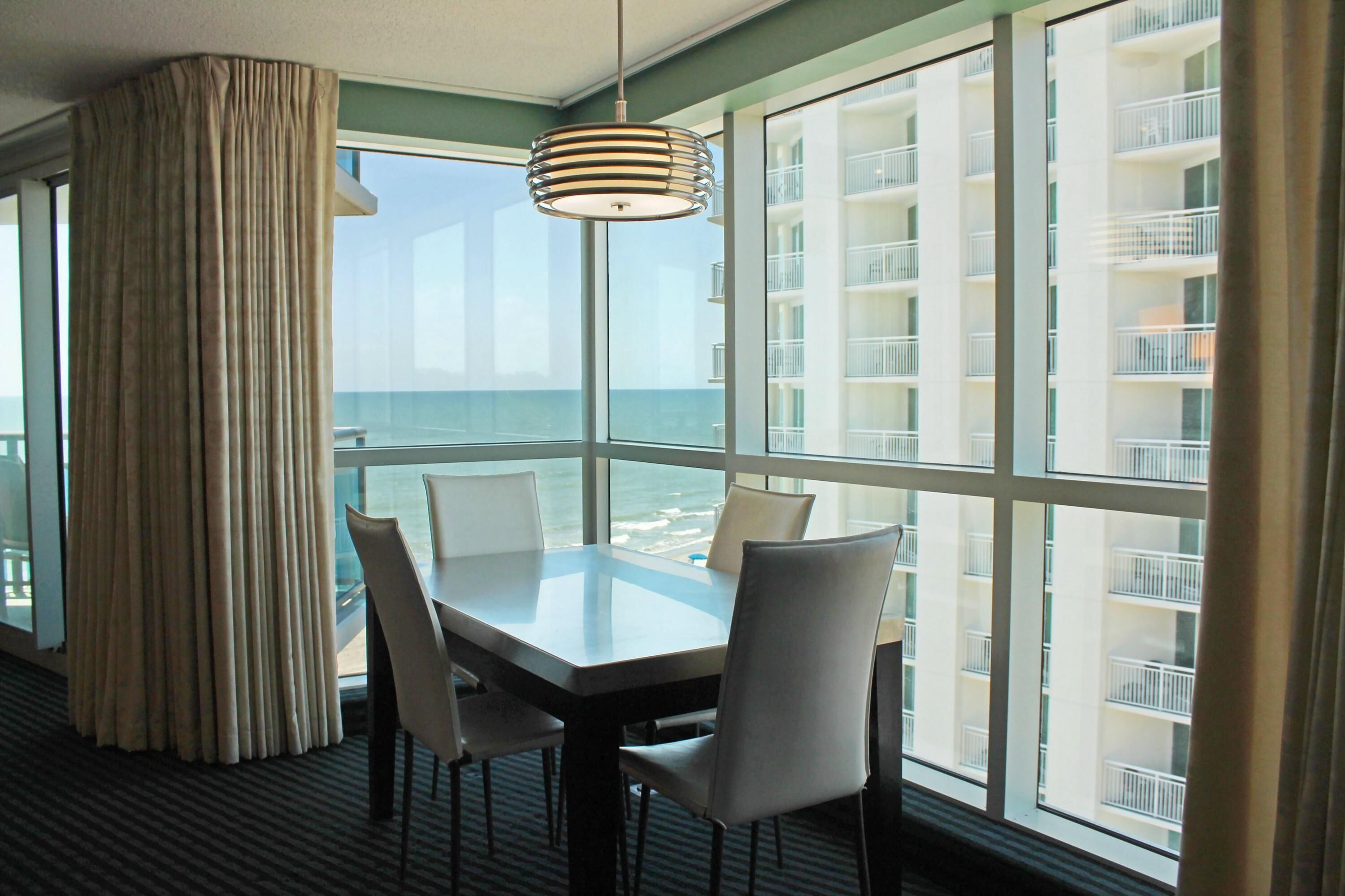 A hotel room with a large window offering a view of the sea and a building with balconies. There is a table with chairs and a hanging light fixture.