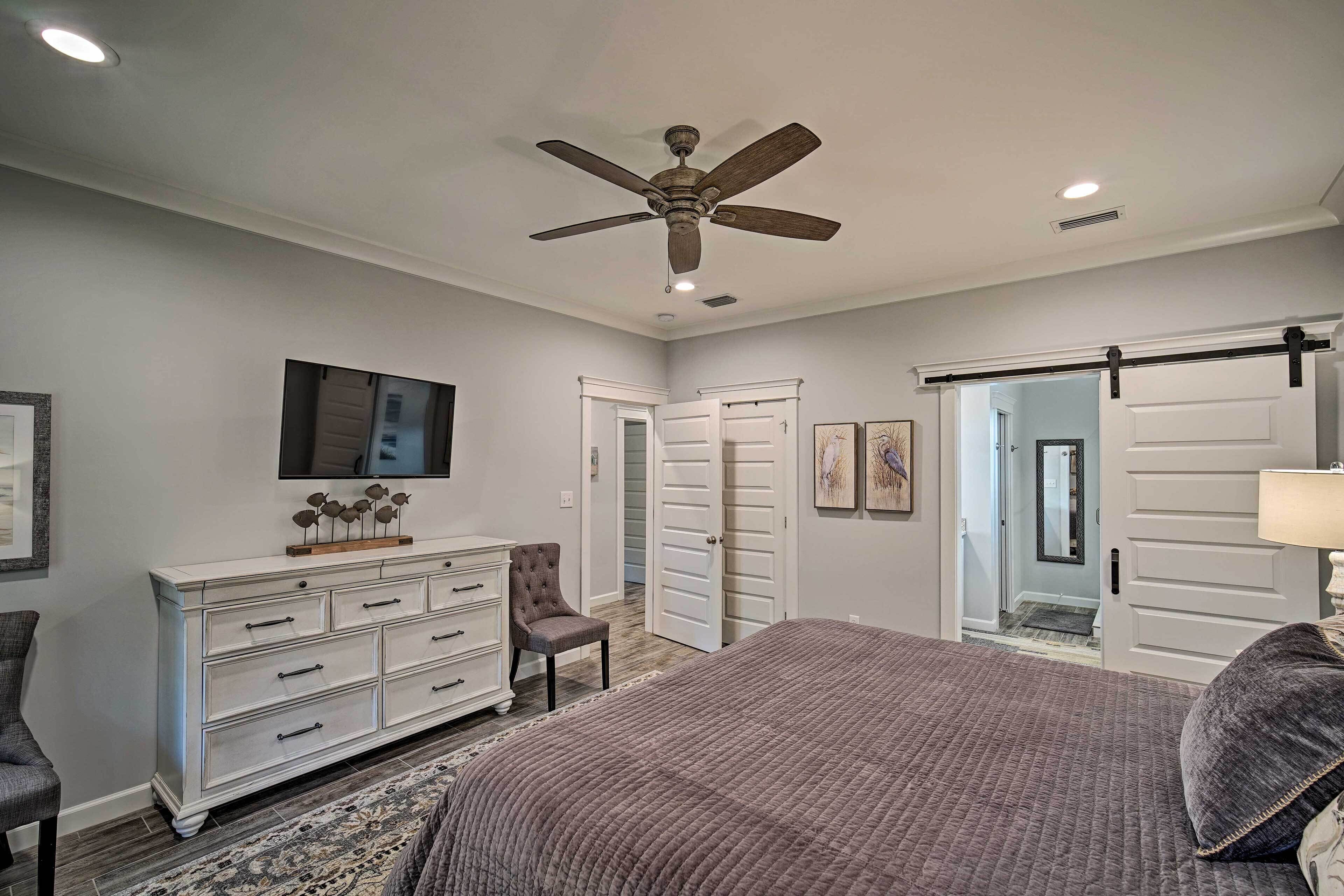 A bedroom with a bed, dresser, chair, ceiling fan, and a door leading to another room.