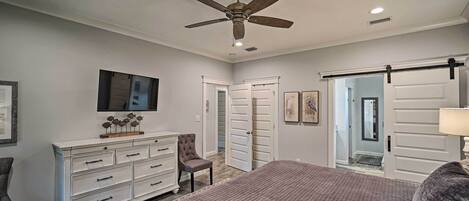 A bedroom with a bed, dresser, chair, ceiling fan, and a door leading to another room.