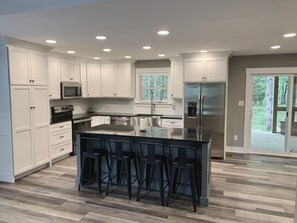 A modern kitchen with white cabinets, a stainless steel refrigerator, a built-in island with bar stools, and a window with blinds.