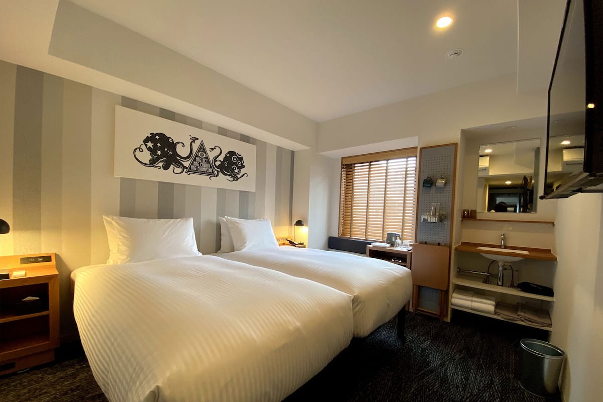 A hotel room with two beds, a large mirror, a sink, and a window with blinds.