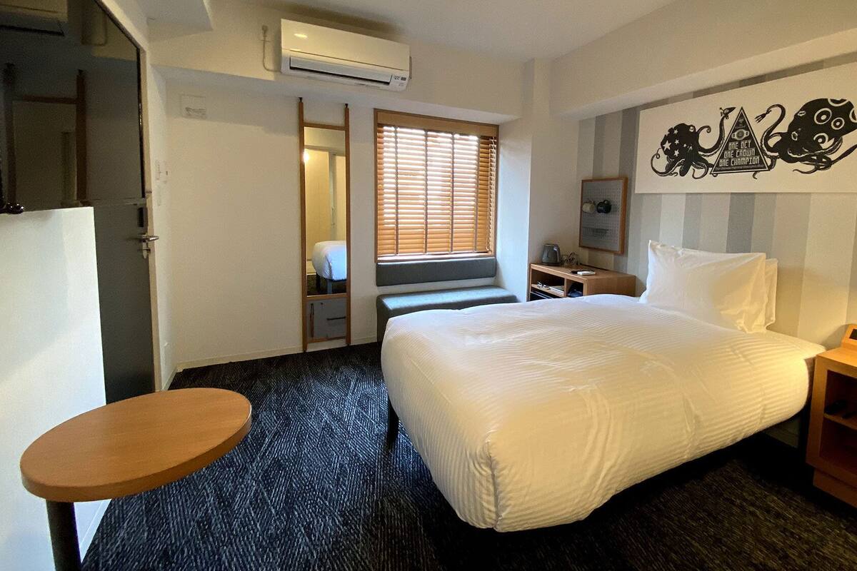 A hotel room with a bed, a desk, a chair, a small table, and a window with blinds.