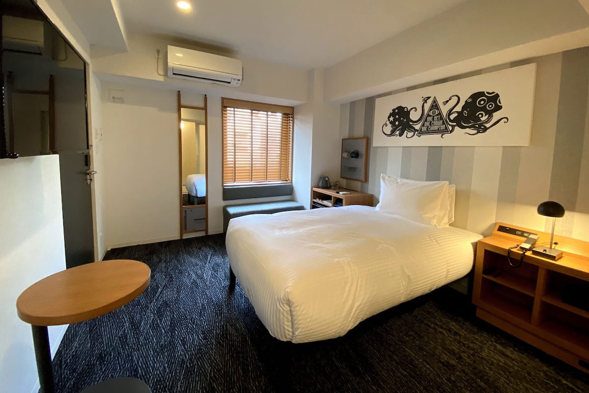 A hotel room with a bed, a desk, a chair, an air conditioning unit, a window with blinds, and a wall decoration.