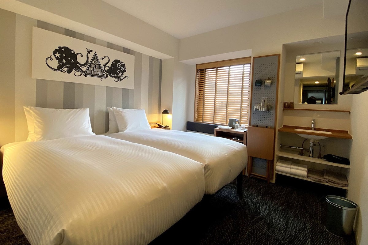 A hotel room with two beds, a desk, a mirror, and a window with blinds.