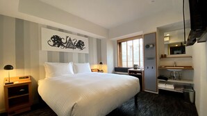 Habitació d'hotel amb un llit gran, una taula de nit amb una làmpada, una finestra amb persianes, una cuina americana petita amb un rentatall i un mirall sobre el rentatall.