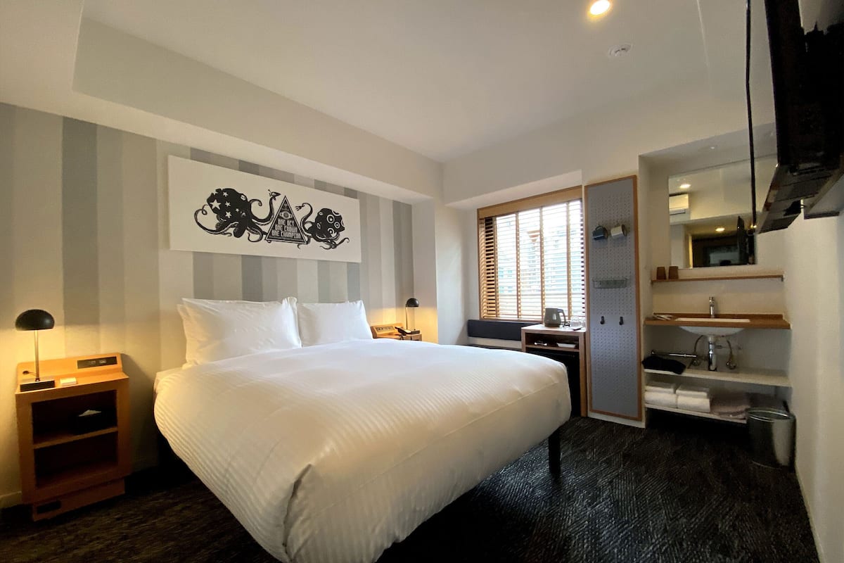 A hotel room with a large bed, a bedside table with a lamp, a window with blinds, a small kitchenette with a sink, and a mirror above the sink.