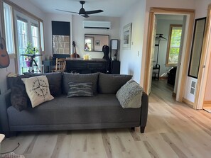 A living room with a grey sofa, a ceiling fan, and a dining area in the background. - Heron's Cove Guesthouse: Peaceful Retreat for Hikes, Antiquing, and Adventures (Stockton Springs)