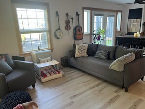 A living room with a grey sofa, a small table, and a window with blinds. - Heron's Cove Guesthouse: Peaceful Retreat for Hikes, Antiquing, and Adventures (Stockton Springs)