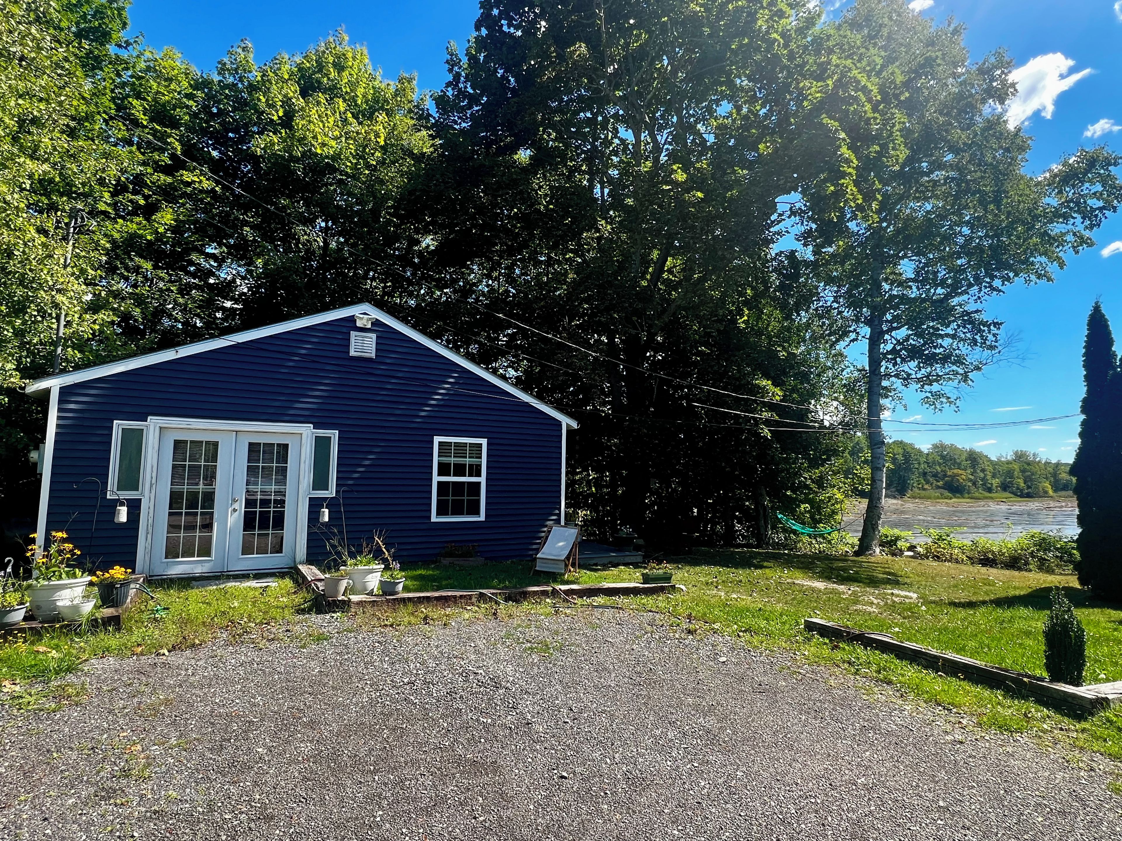 A single-story blue house with a front porch, surrounded by trees and a gravel driveway.