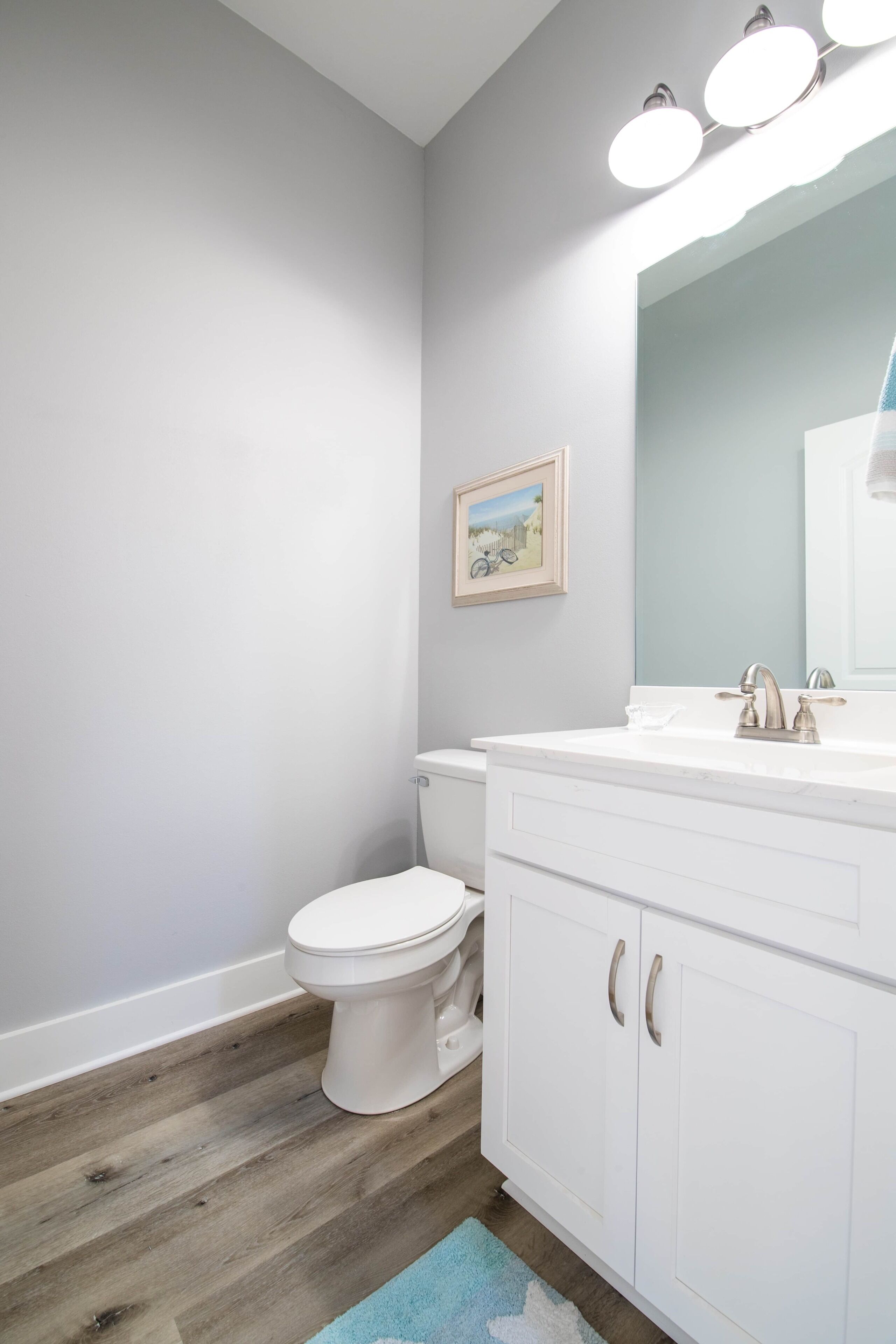 A bathroom with a white toilet, a white vanity with a mirror, and a light blue rug.