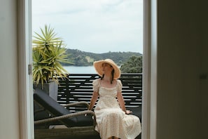 A person sitting on a wicker chair on a balcony with a view of a lake and hills. - Black Bach on Burrell (Oneroa)
