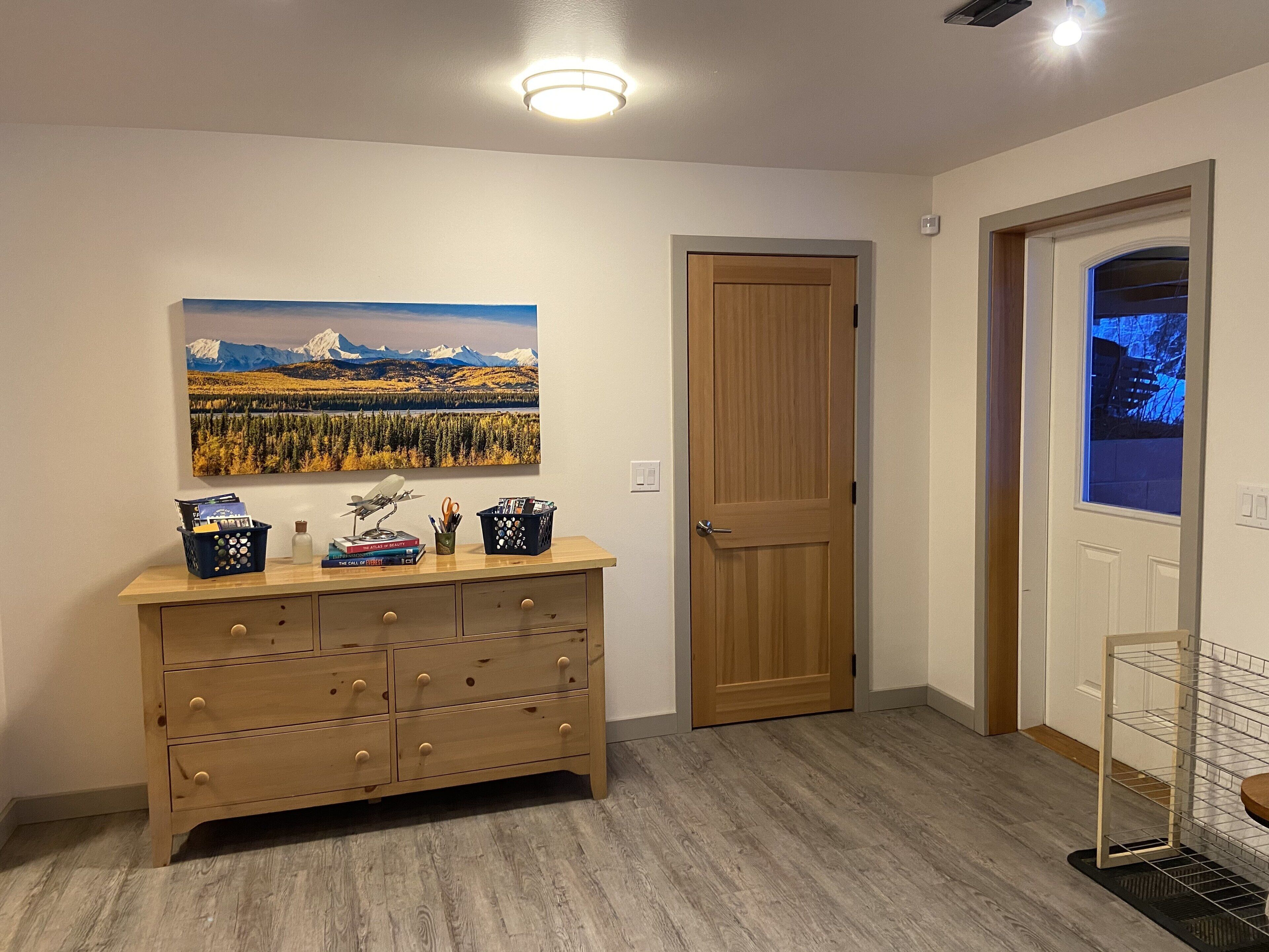 A wooden dresser with drawers, a framed landscape painting, a door, and a window with a view.