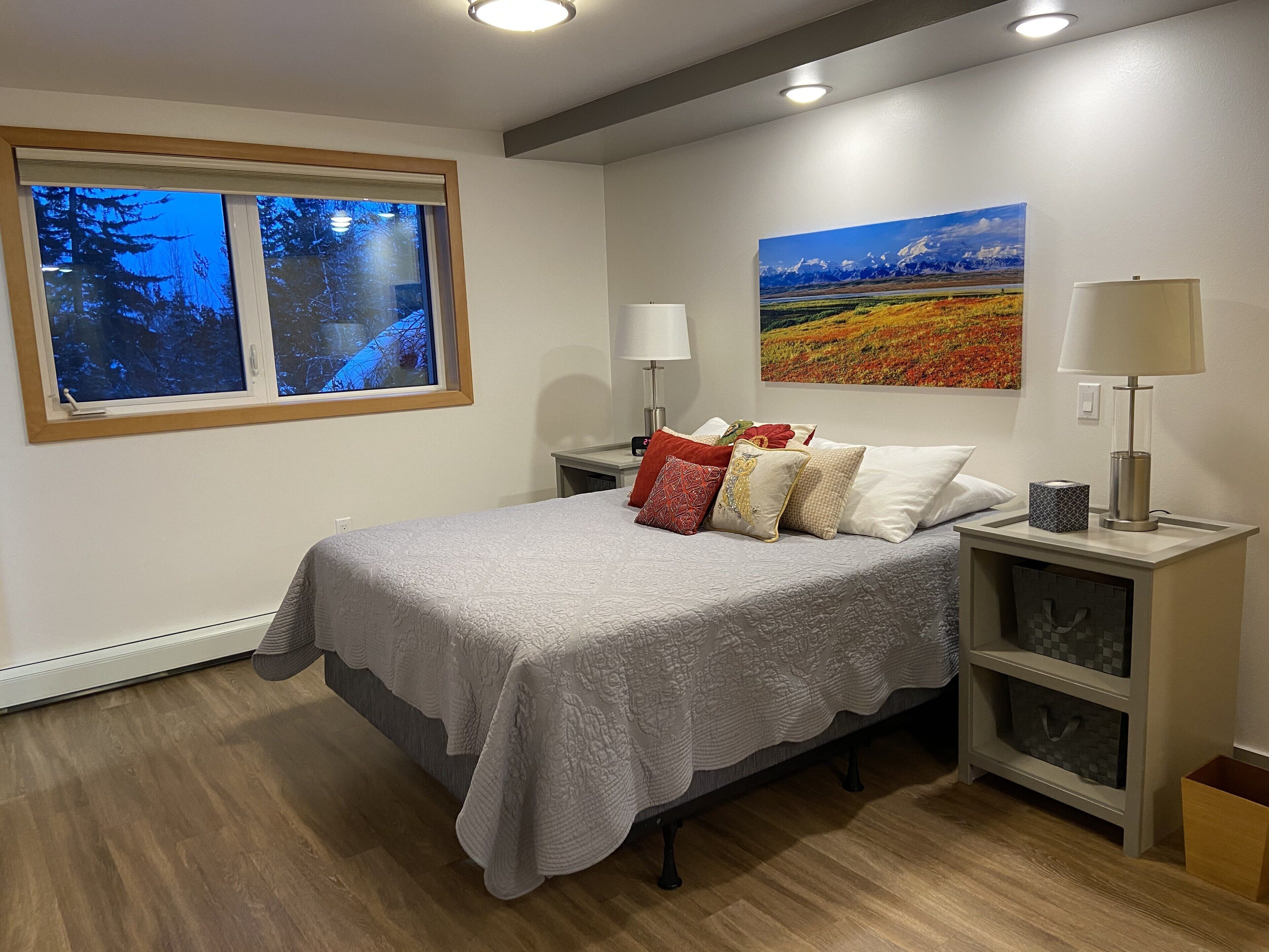 Un dormitorio con cama, mesita de noche, lámpara, un cuadro y ventana con vista a árboles.