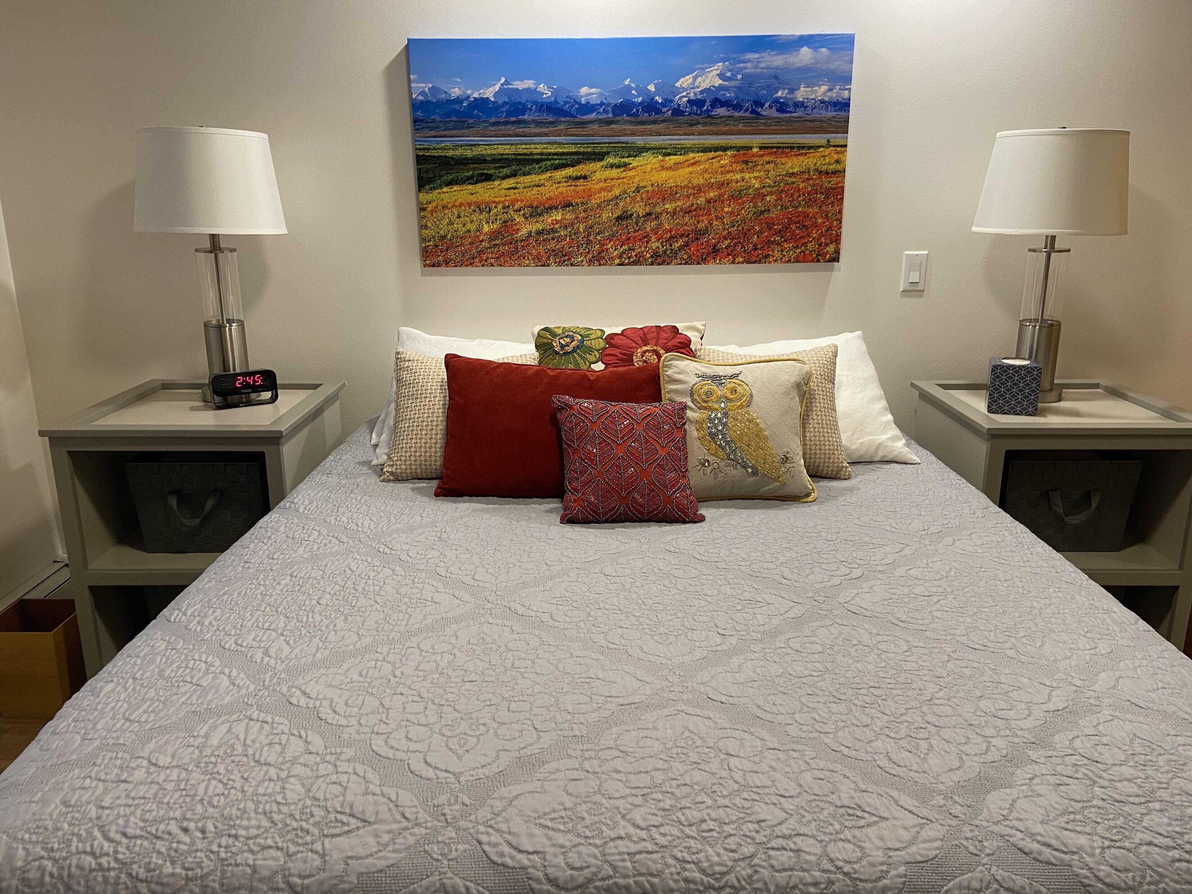 Una cama bien hecha con un patrón acolchado, flanqueada por dos mesitas de noche con lámparas y un reloj digital. Un cuadro de paisaje grande está centrado sobre la cama.
