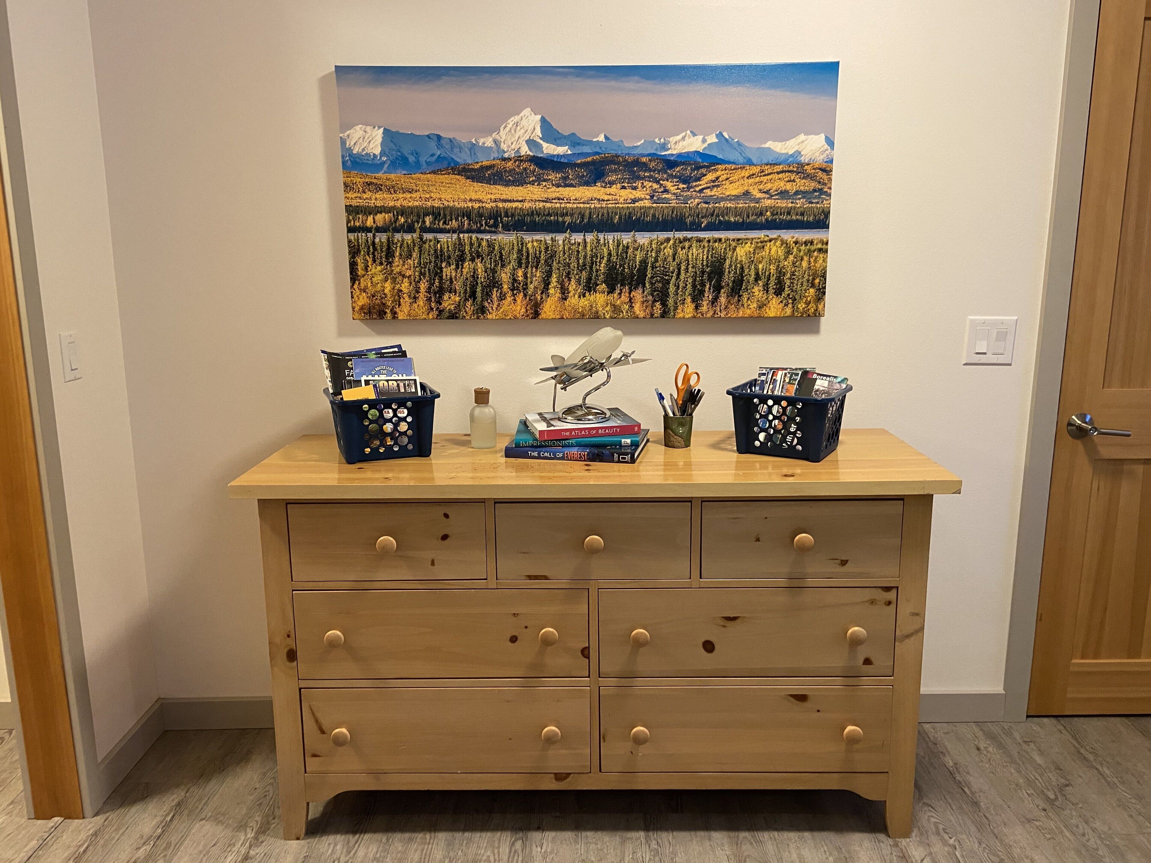 Une commode en bois avec six tiroirs, un tableau représentant un paysage montagneux et deux paniers contenant des livres et du matériel de bureau.