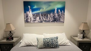 Hotellihuone, jossa on sänky, kaksi yöpöytää, digitaalinen kello, revontulikuva ja kaksi lamppua.