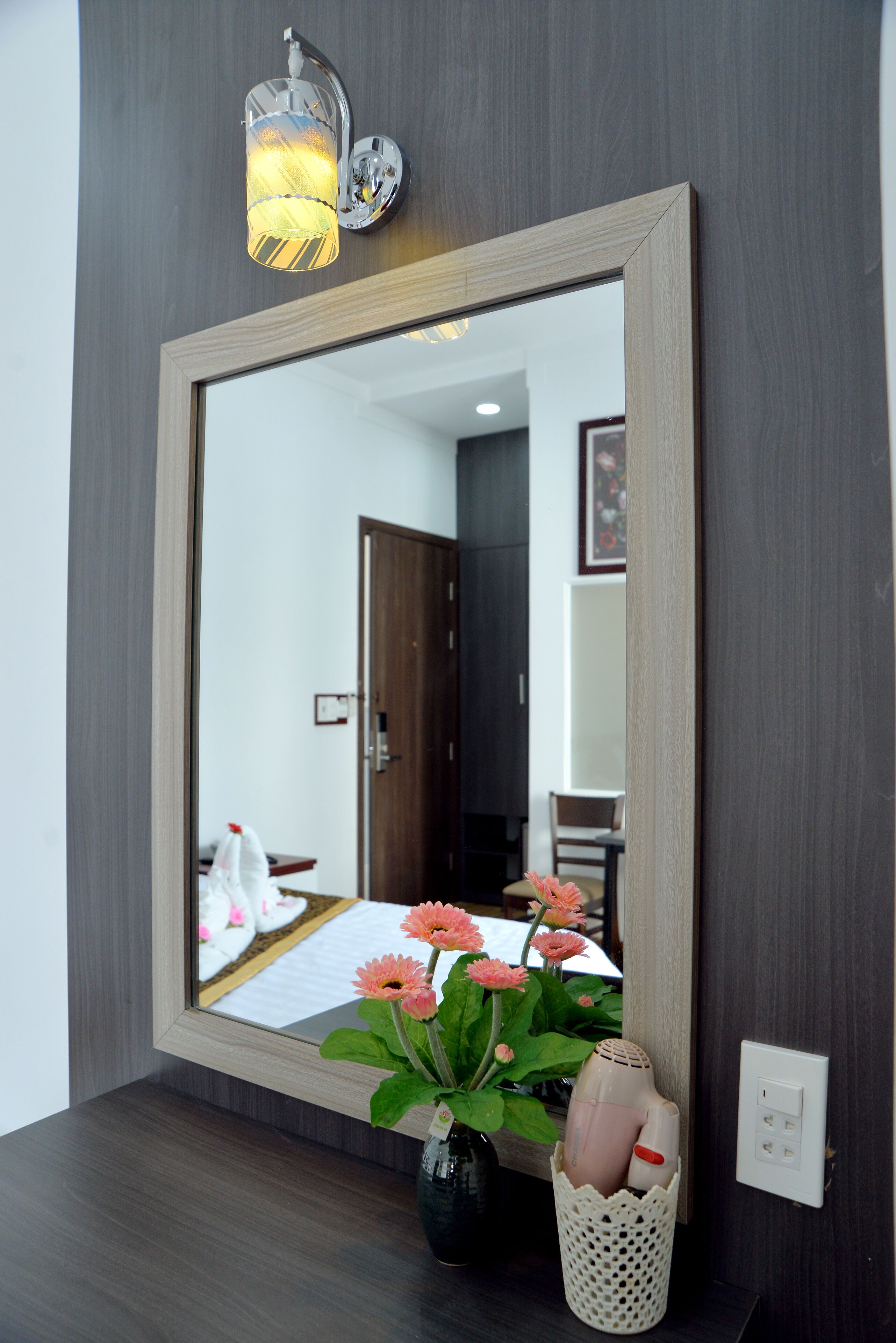 A mirror with a wooden frame, a vase with flowers, and a wall-mounted light fixture.