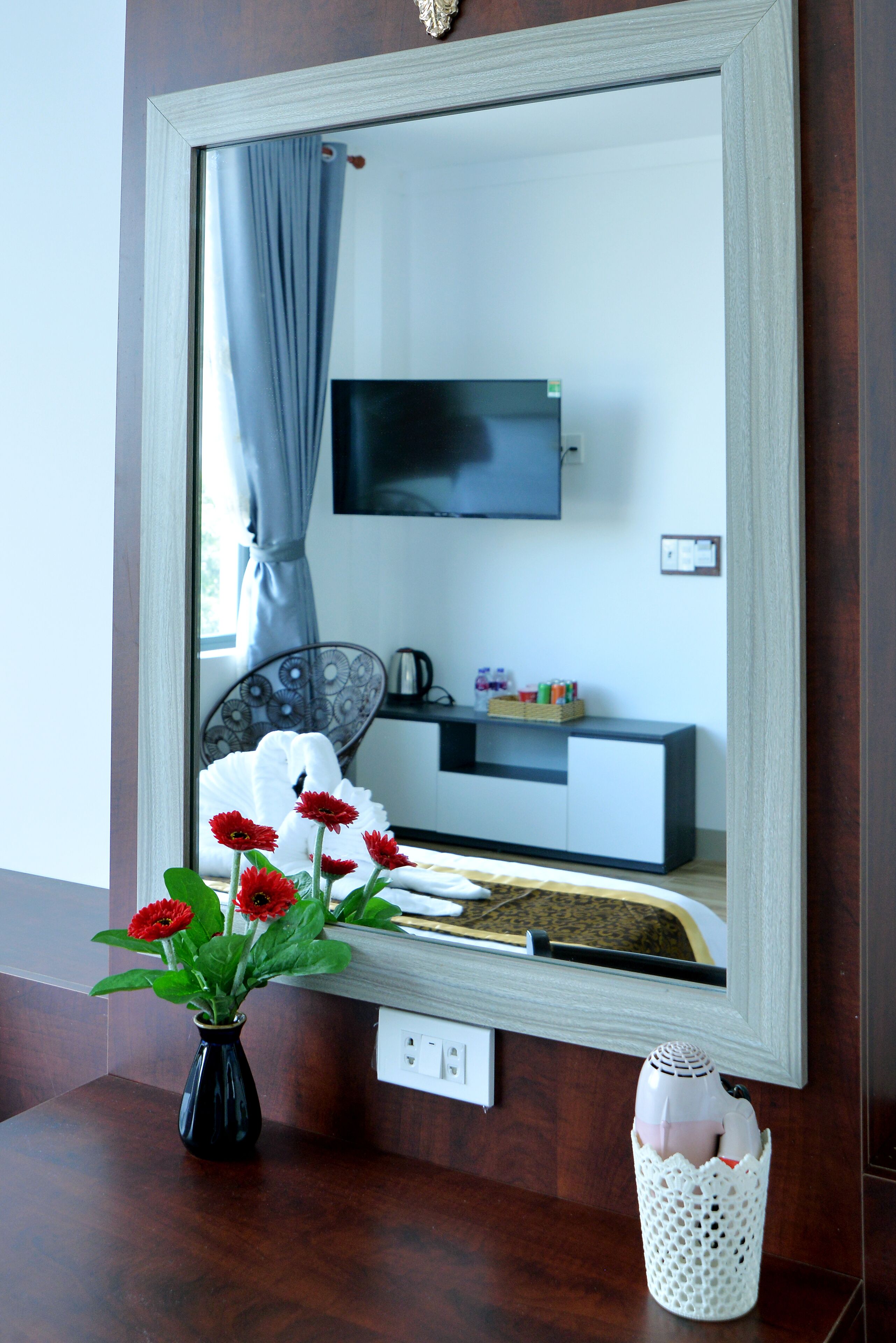 A mirror reflecting a room with a TV, a white cabinet, and a vase with red flowers on a wooden surface.