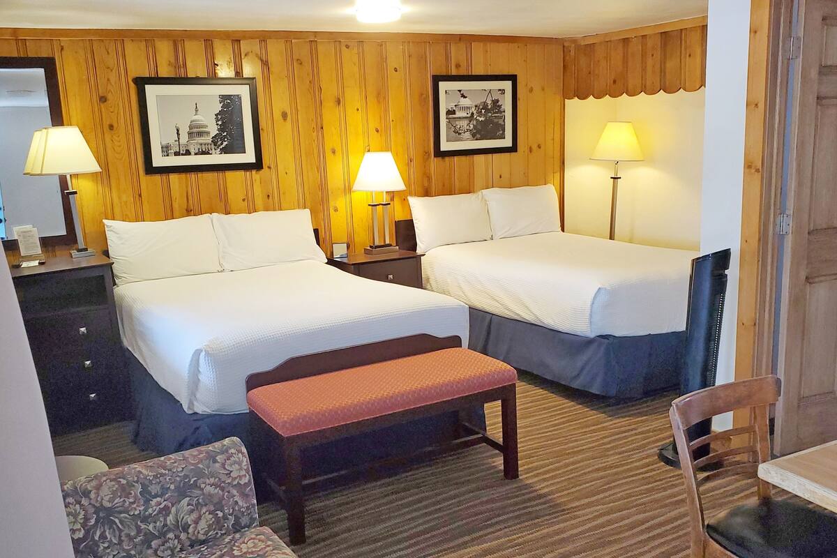 A hotel room with two beds, a wooden headboard, a small table, a chair, and two lamps.