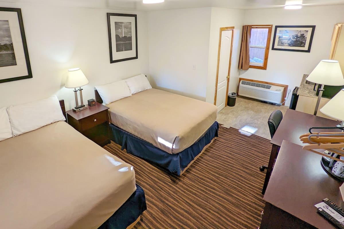 A hotel room with two beds, a desk, a chair, and a small table. There are framed pictures on the walls and a window with curtains.