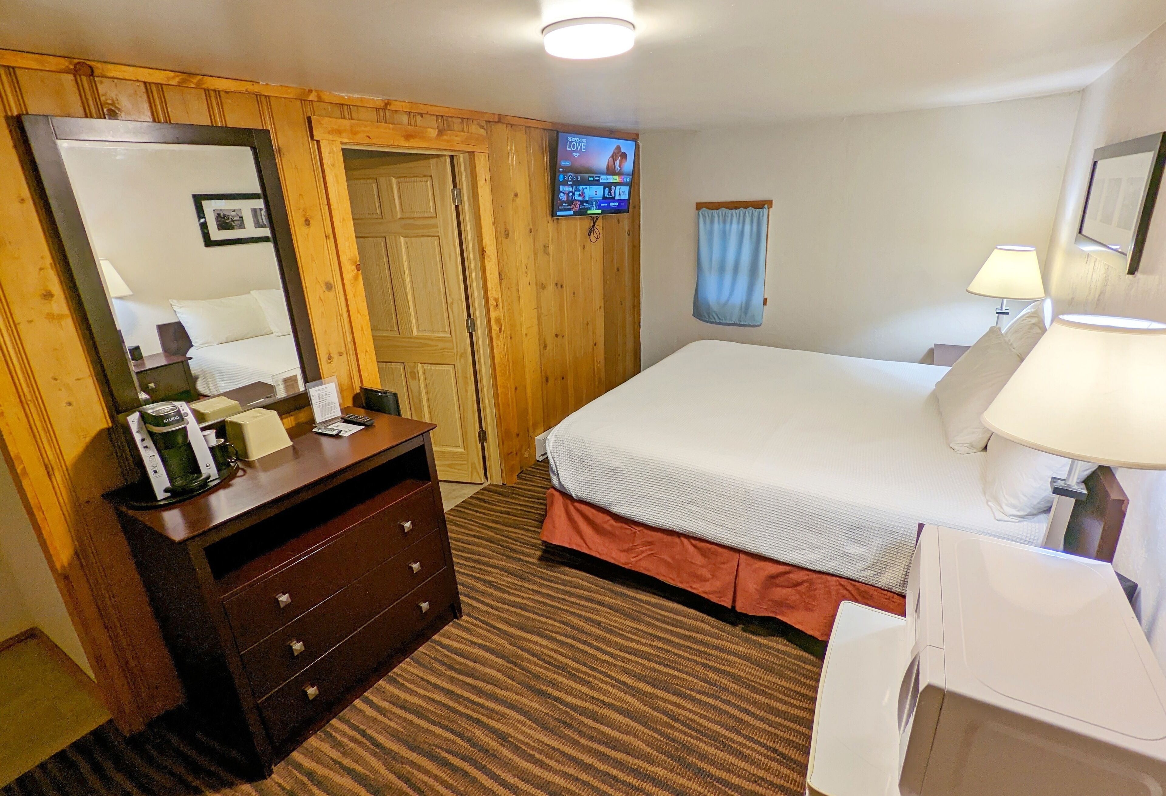 Ein Hotelzimmer mit Bett, Kommode, Spiegel, Fernseher und einem Fenster mit Vorhängen.