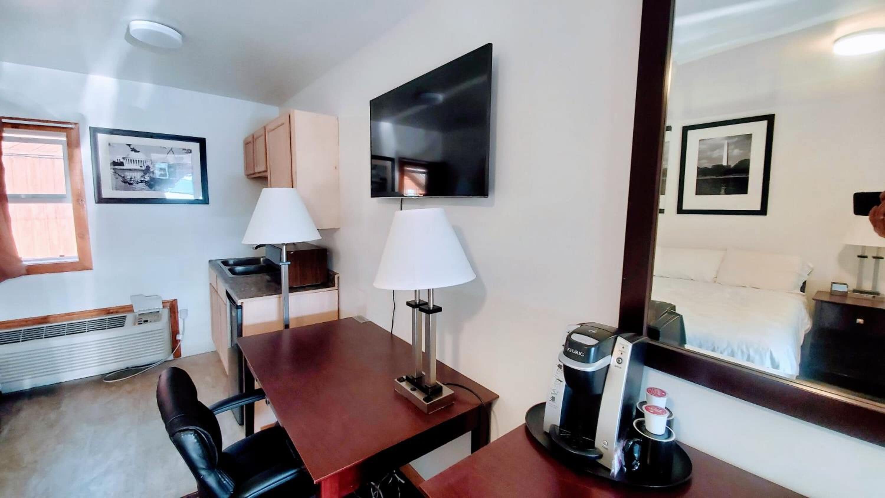 A hotel room with a wooden desk, two lamps, a coffee maker, a bed, and a large mirror.
