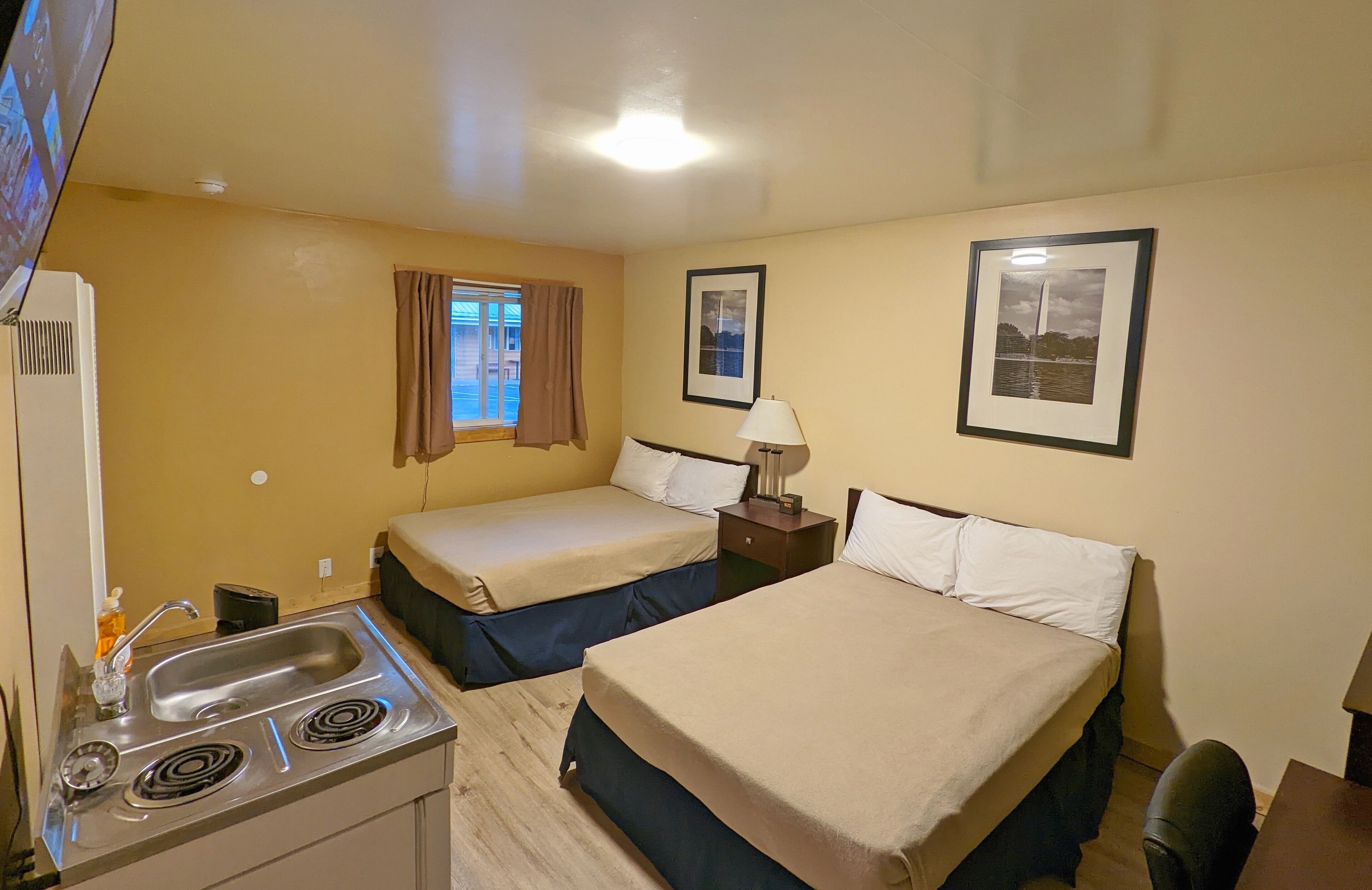 Habitación de hotel con dos camas, un kitchenette, una ventana con cortinas y cuadros en las paredes.
