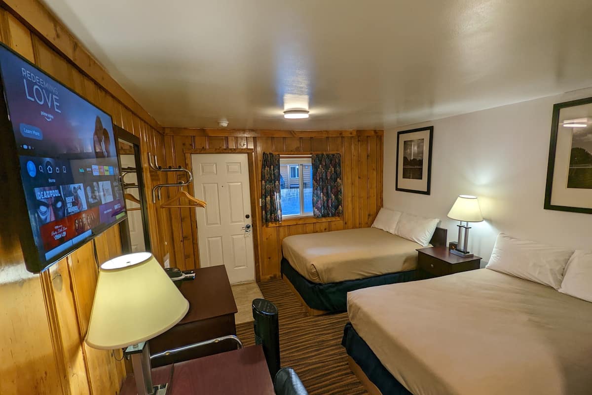 A hotel room with wooden paneling, a TV on the wall, two beds, a desk, and a window with curtains.