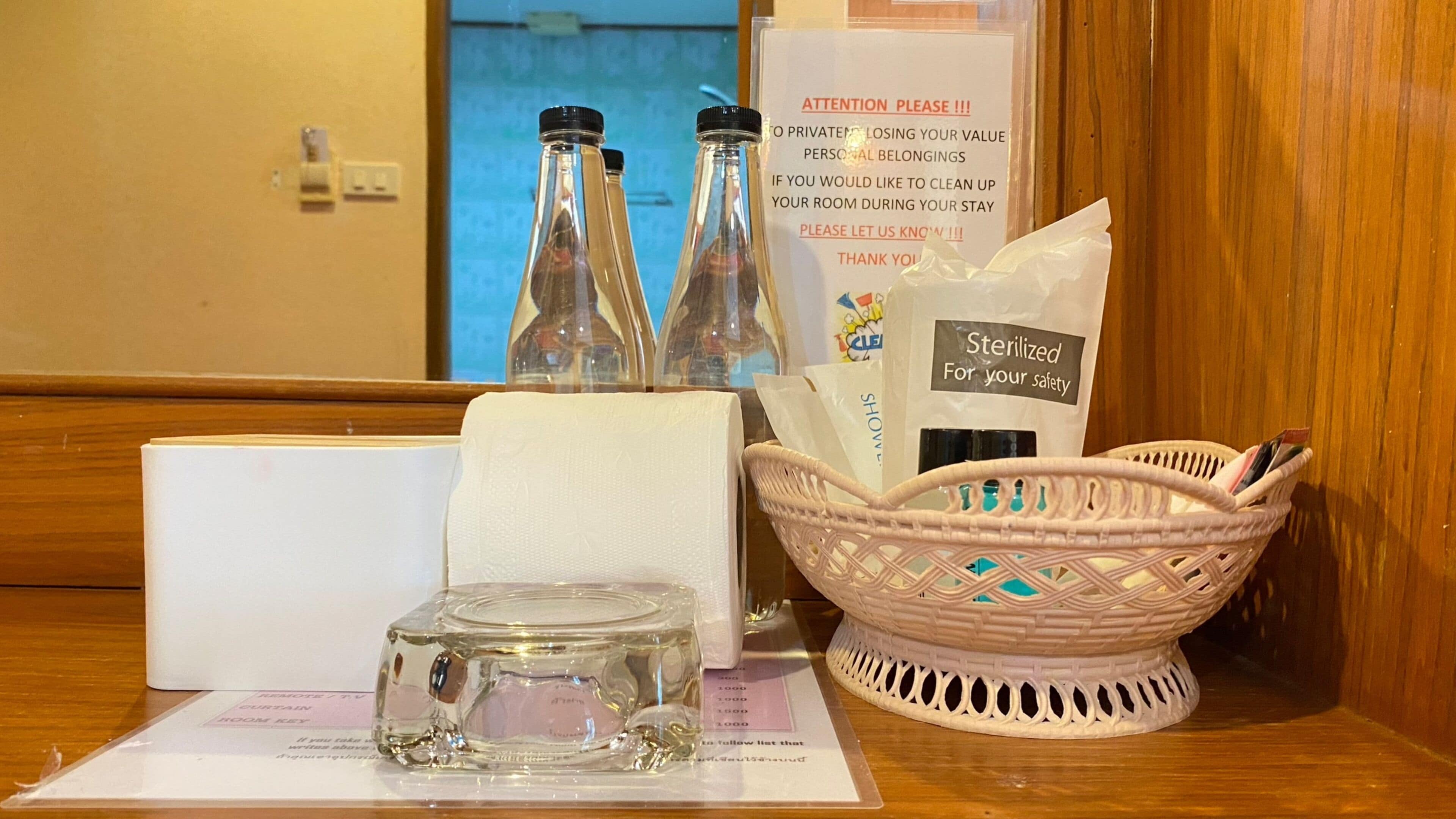 Zona de recepción de una habitación de hotel, con una botella de vidrio, una caja de pañuelos, una cesta con artículos de aseo y un cartel sobre limpieza.