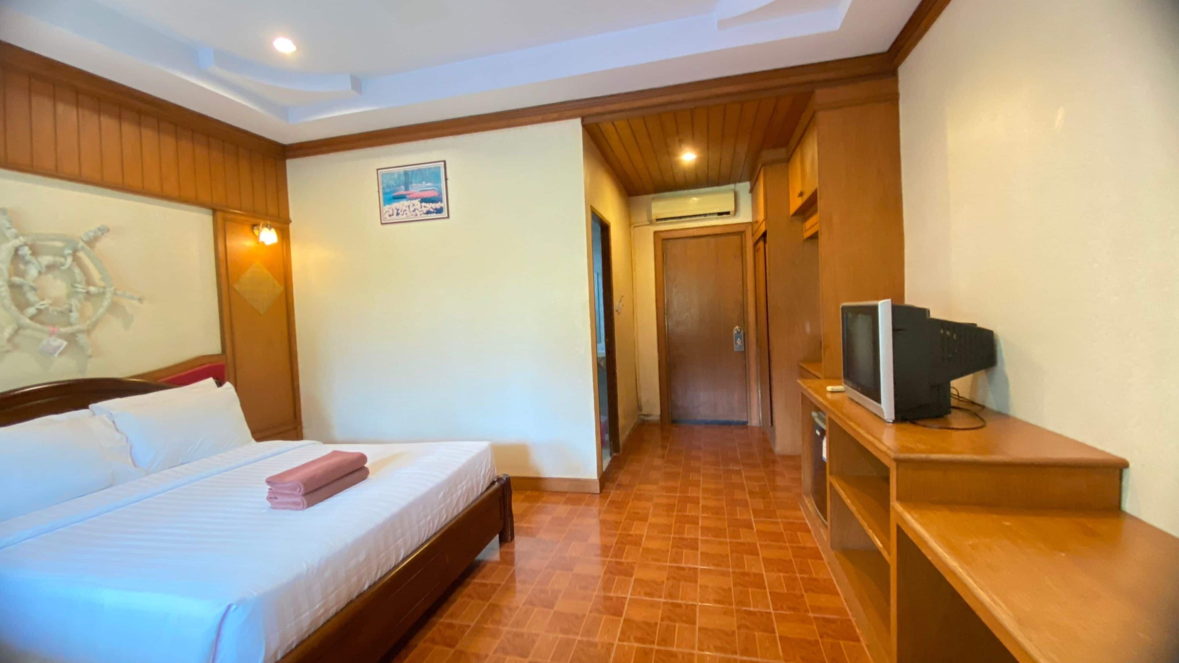 Habitación de hotel con una cama, un televisor sobre un mueble de madera y un pasillo que lleva a una puerta.