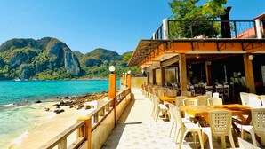 A beachfront restaurant with outdoor seating, a view of the sea, and mountainous terrain in the background.