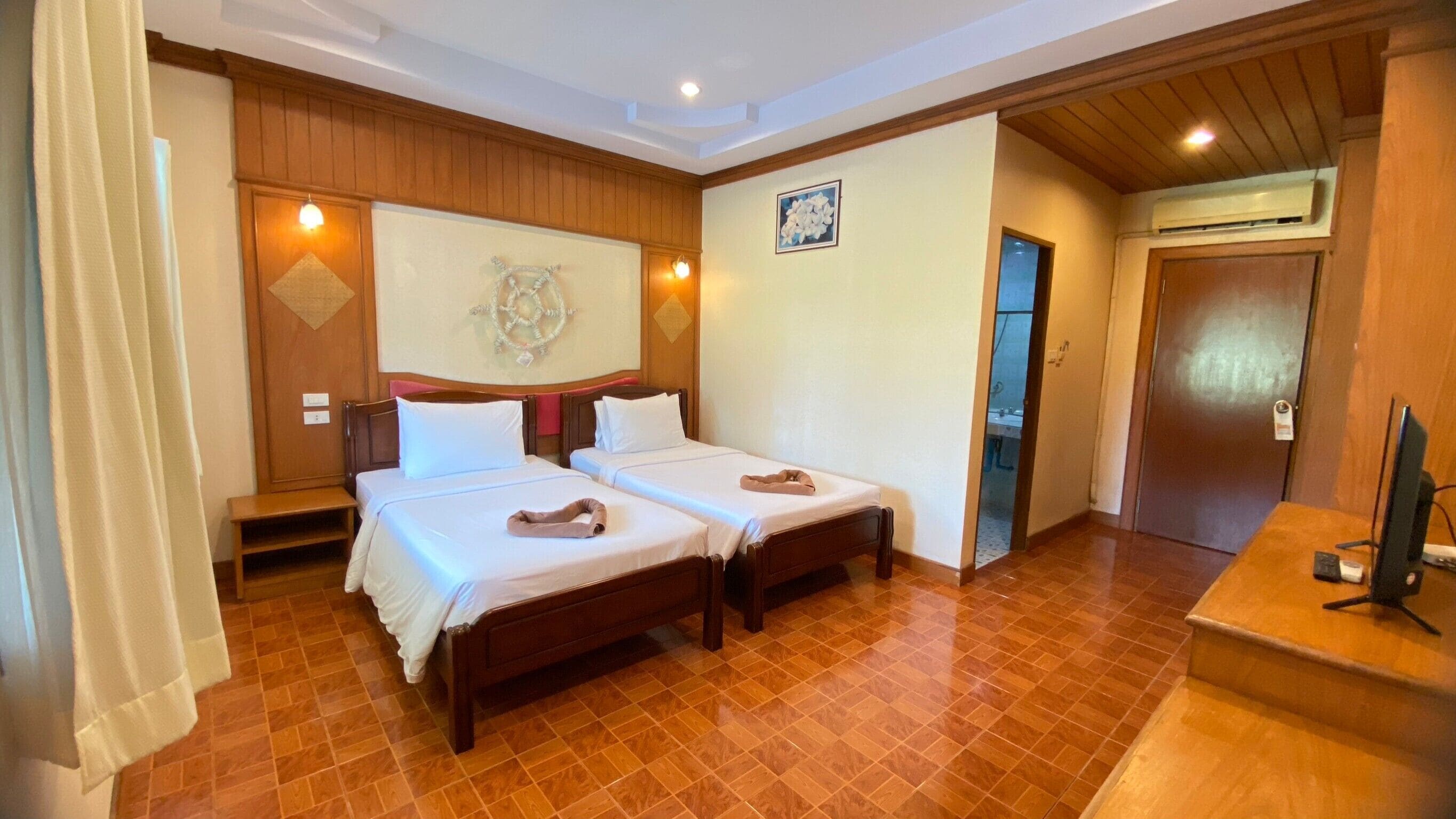 A hotel room with two beds, wooden flooring, a desk with a computer, and a door leading to a bathroom.