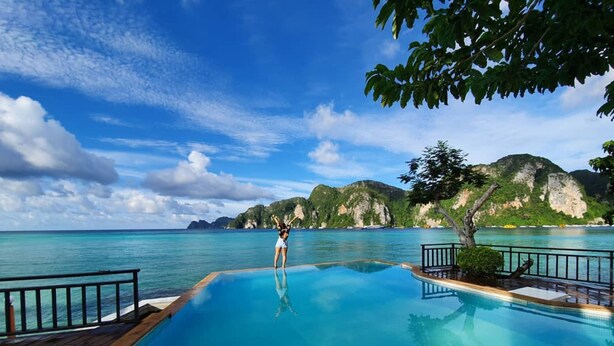 A pool with clear blue water, surrounded by a wooden deck and metal railings. In the background, there are green hills and a clear sky.