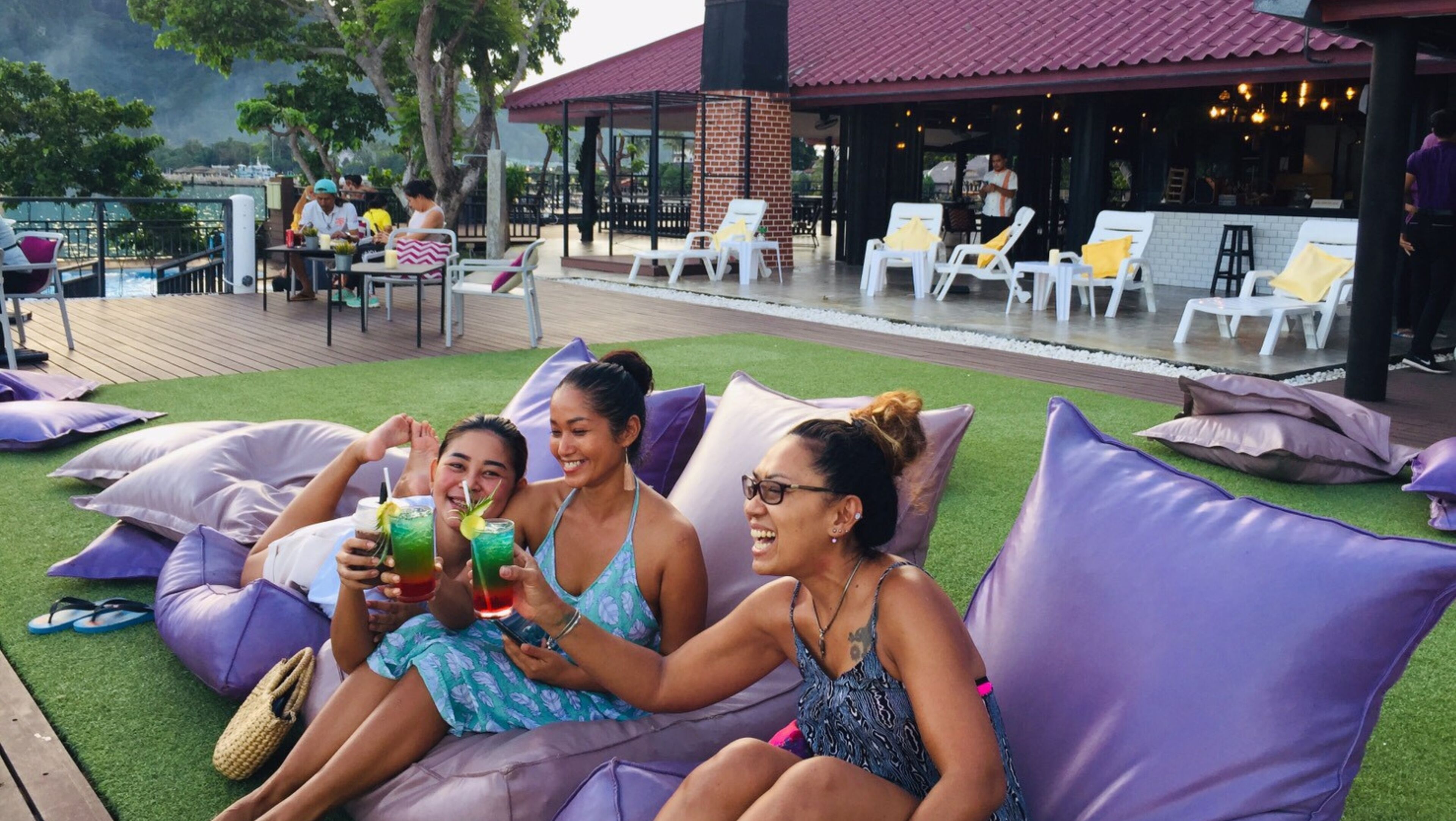 A group of people relaxing on large purple cushions on a grassy deck with a pool in the background.