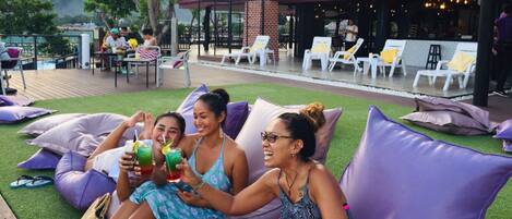 A group of people relaxing on large purple cushions on a grassy deck with a pool in the background.