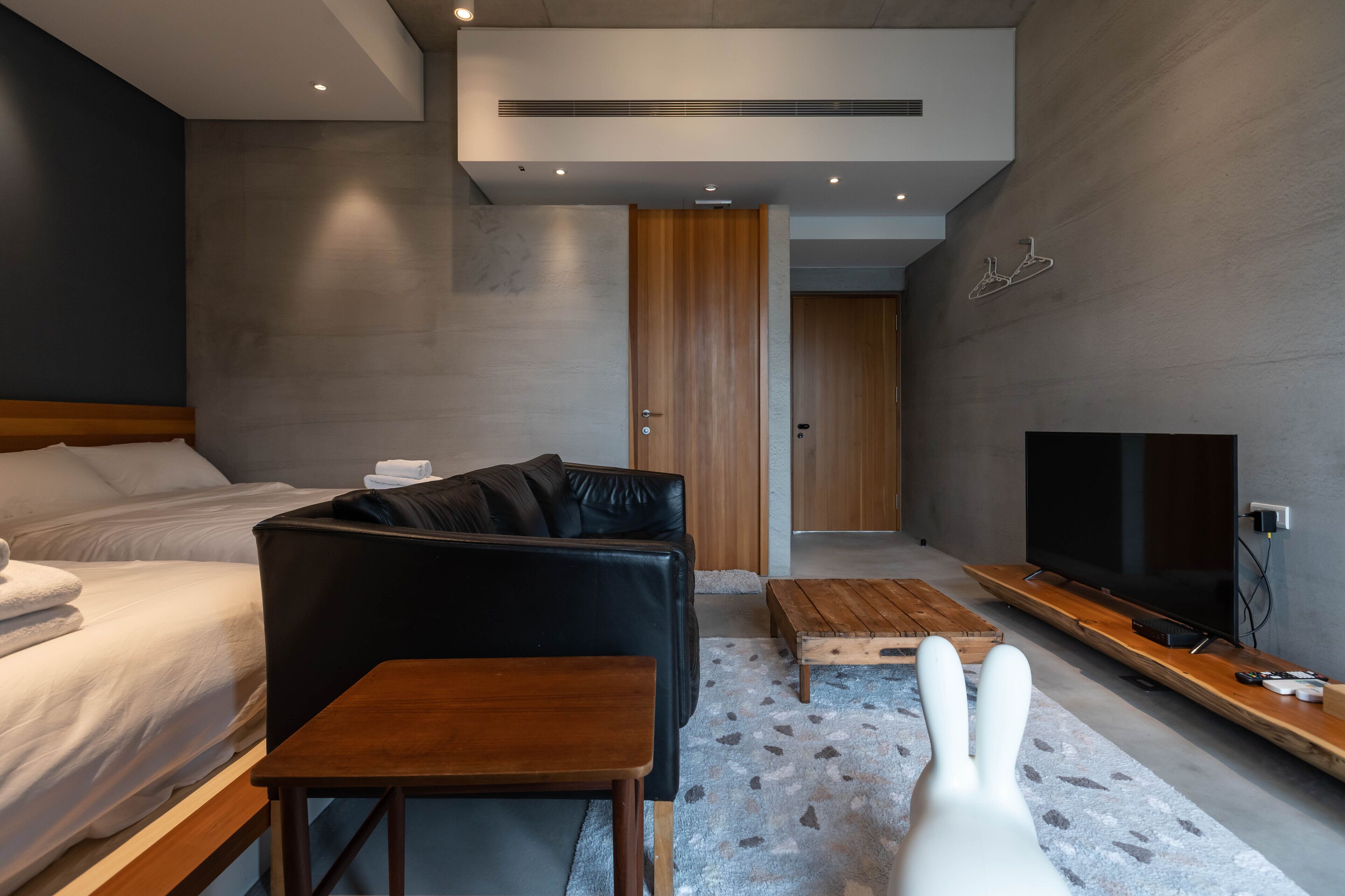 A modern hotel room with a bed, a black leather sofa, a wooden coffee table, a TV, and a white chair.
