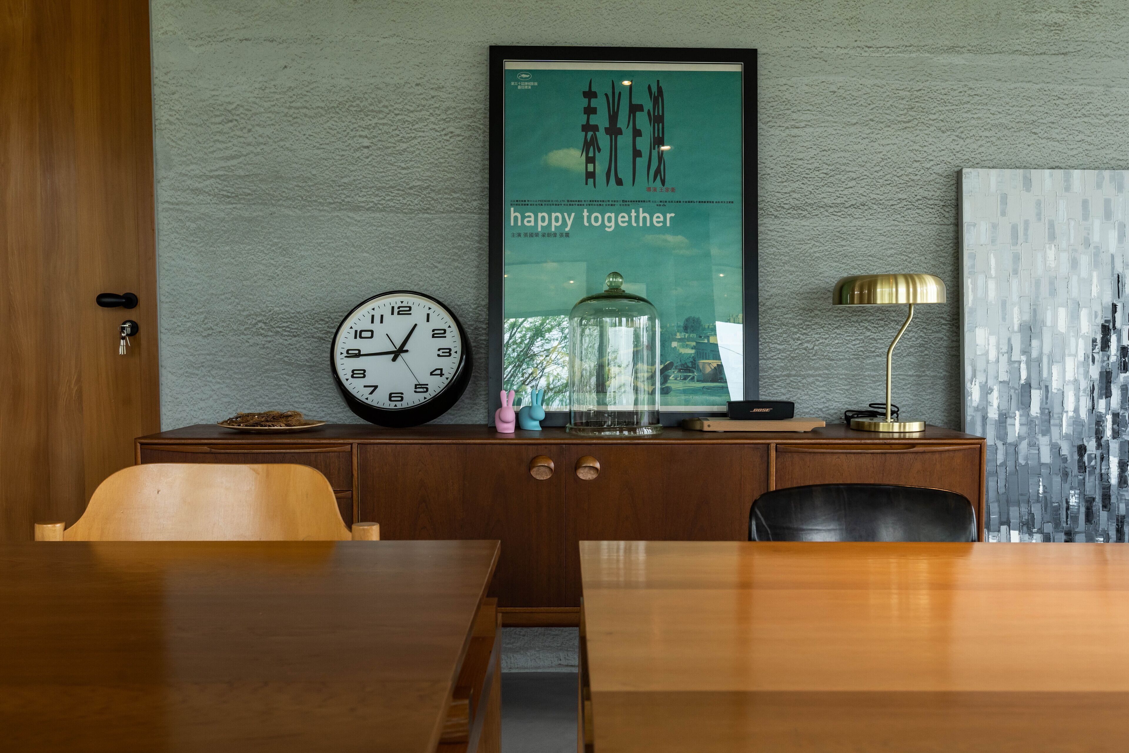 A dining area with a wooden table and chairs, a clock, a decorative item on the wall, and a framed picture with text.