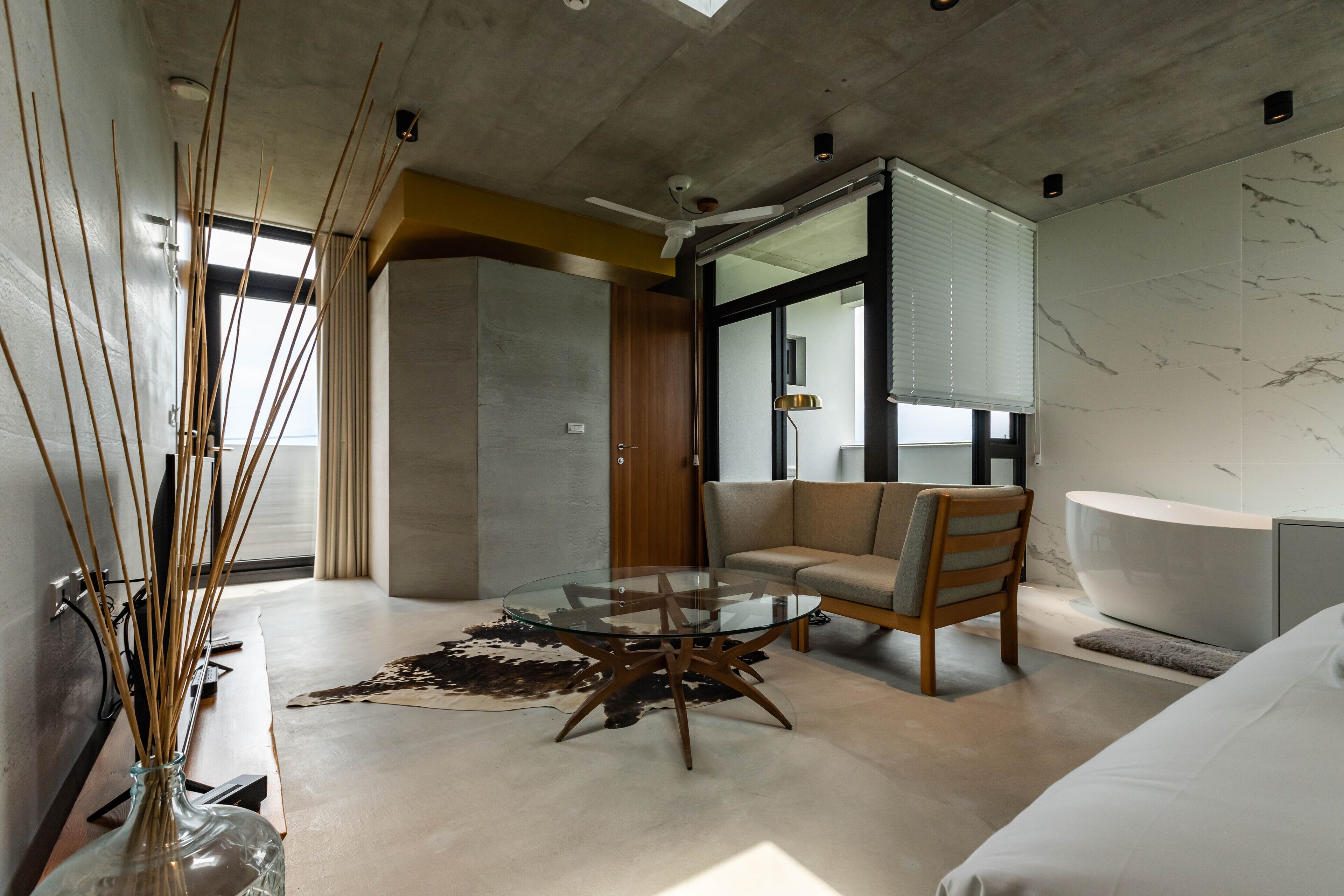 A modern hotel room with a glass coffee table, a sofa, a bathtub, and a large window with blinds.