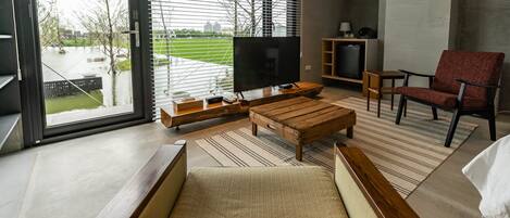 Una sala de estar moderna con un televisor grande, muebles de madera y vistas a un parque a través de amplios ventanales.