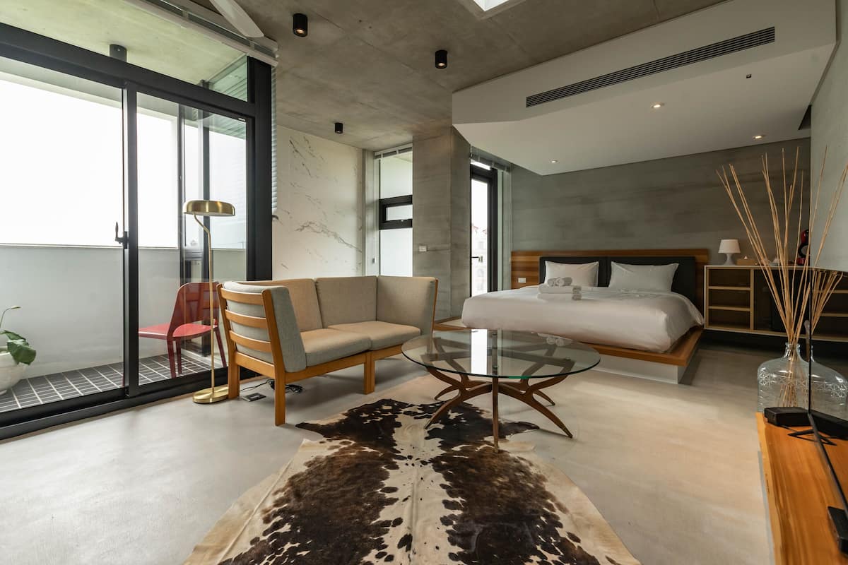 A modern hotel room with a glass door leading to a balcony, a glass coffee table, a sofa, a bed with a wooden headboard, and a large cowhide rug.